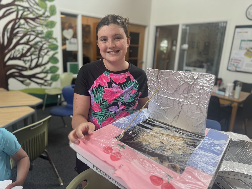 A young person displays a science project with foil and a colorful design. Background includes desks, chairs, and a tree mural.