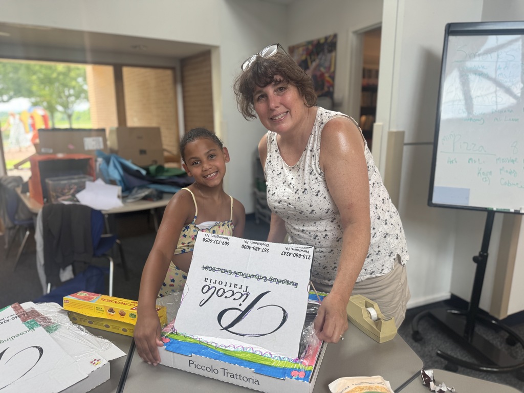 Woman and child hold a box labeled Books. A desk is behind them with a whiteboard on a stand.