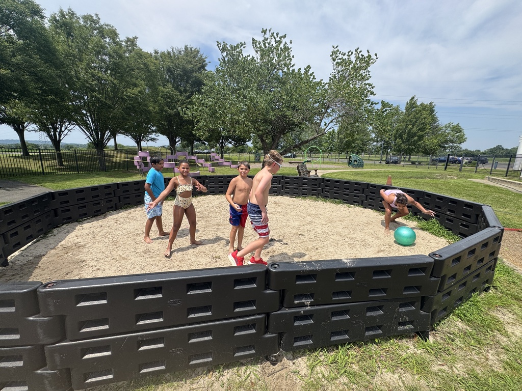 A group of people play beach volleyball in a sand pit surrounded by black barriers. Trees are visible in the background.