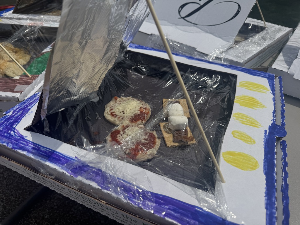 Pizza, crackers, and marshmallows are on a black tray under a plastic sheet in a blue-bordered box.