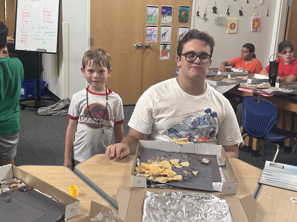 Two boys in a classroom, one wearing glasses, standing at a table with a pizza box.