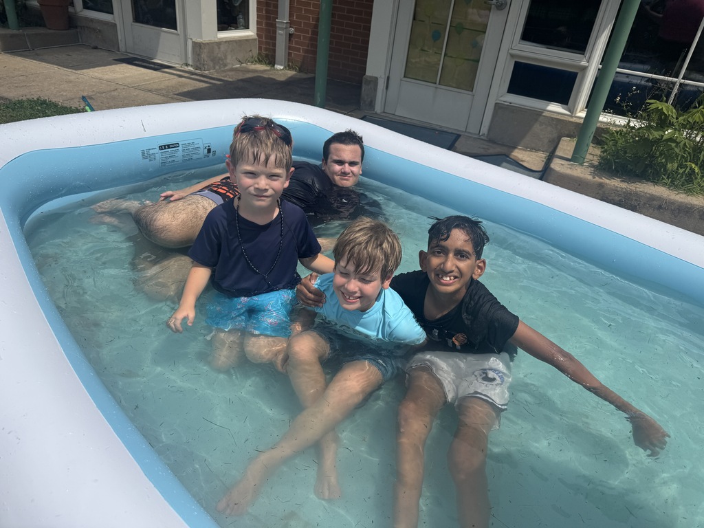 Four children in swim attire in a small pool with a building in the background.