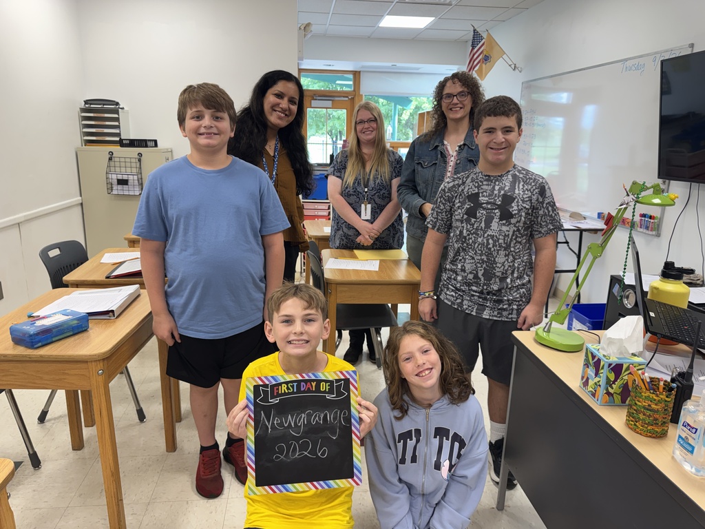 Group of students and adults in a classroom, some standing, others seated. Boy holds a sign, and desks and a whiteboard are visible.