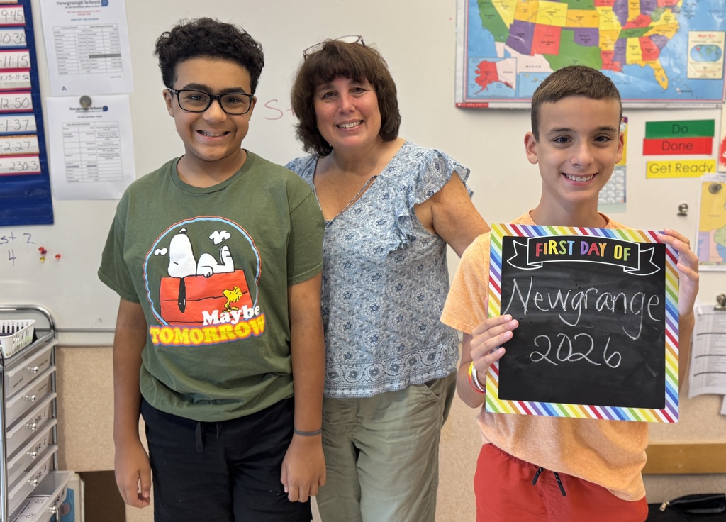 Two boys and a woman stand in a classroom. One boy holds a sign saying "Newgrange 2026".