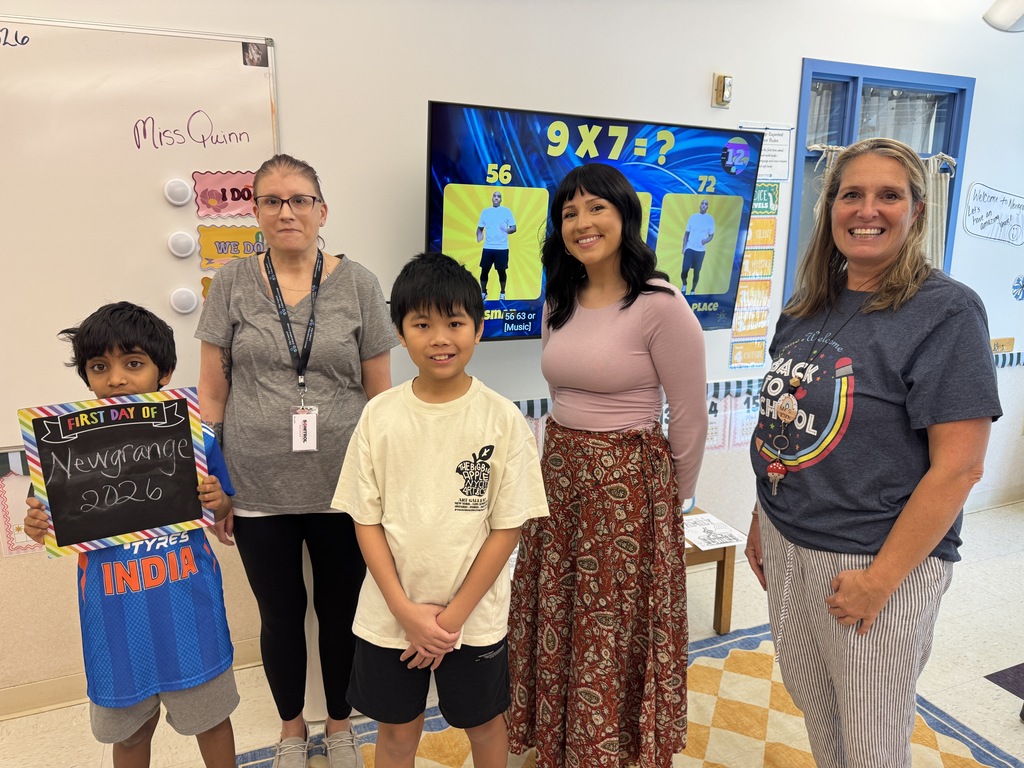 Four people in a classroom. Two boys stand in front, one holding a sign. Two women stand behind them. A large screen displays numbers.