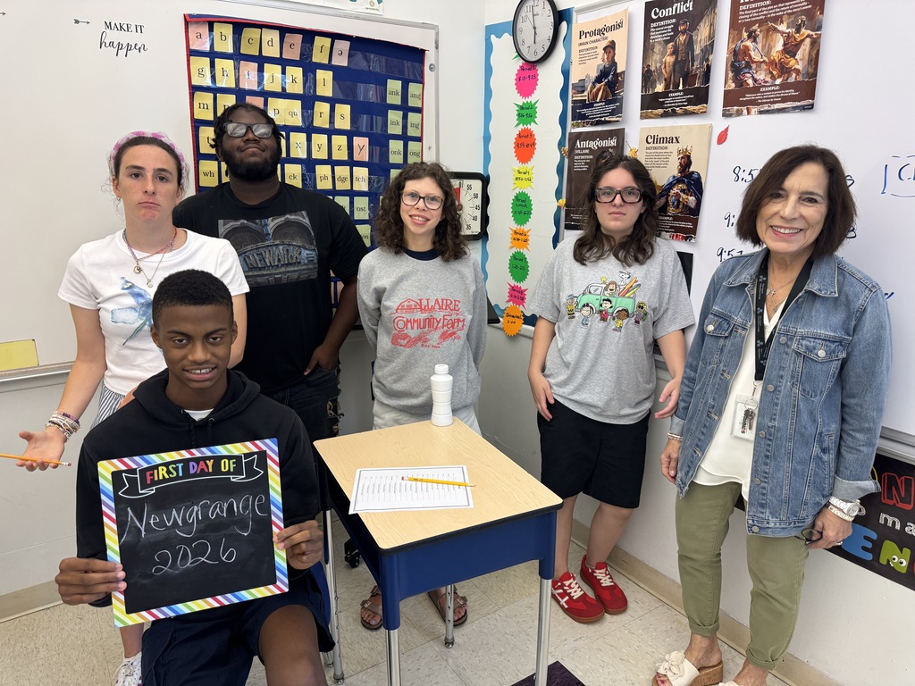 Several people pose for a picture in a classroom with a teacher. One person holds a sign reading "Newgrads 2026."