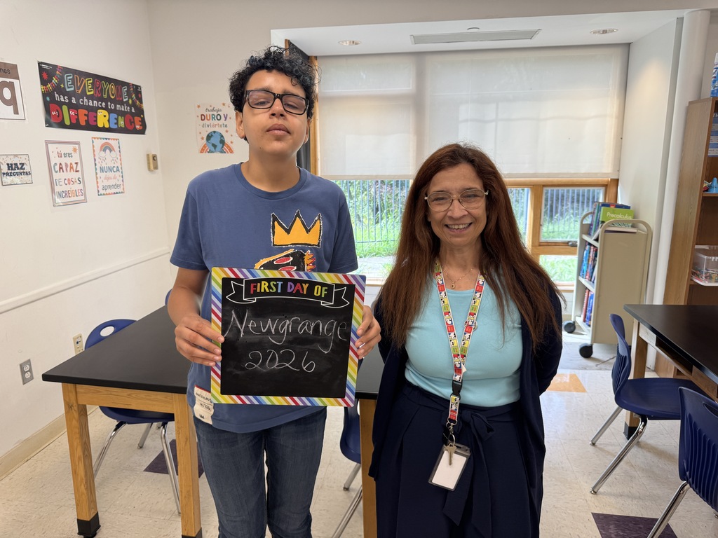 A boy in a blue shirt and glasses holds a sign. A woman with glasses and a lanyard stands next to him. Behind them, tables, chairs, and a whiteboard are visible.