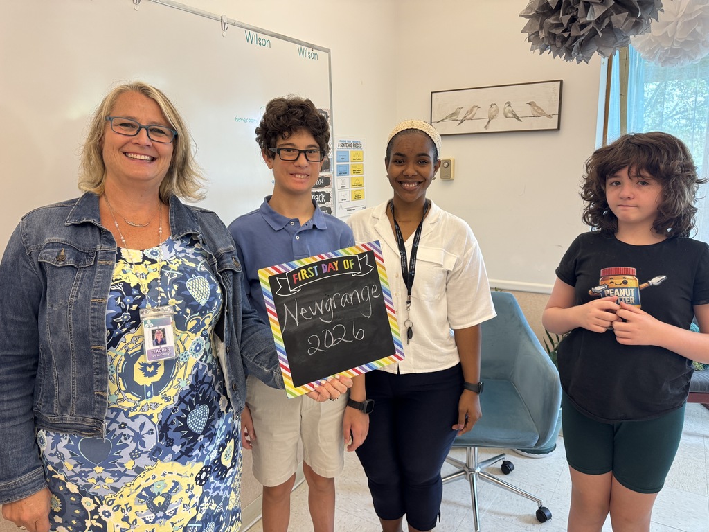 Four people in a room, two girls and a woman, standing close. A woman holds a blackboard with text.