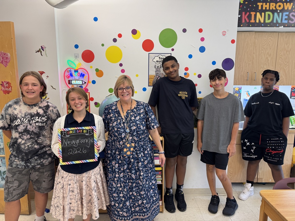 A group of young people and a woman stand in a room with a colorful wall of dots. They hold a sign.