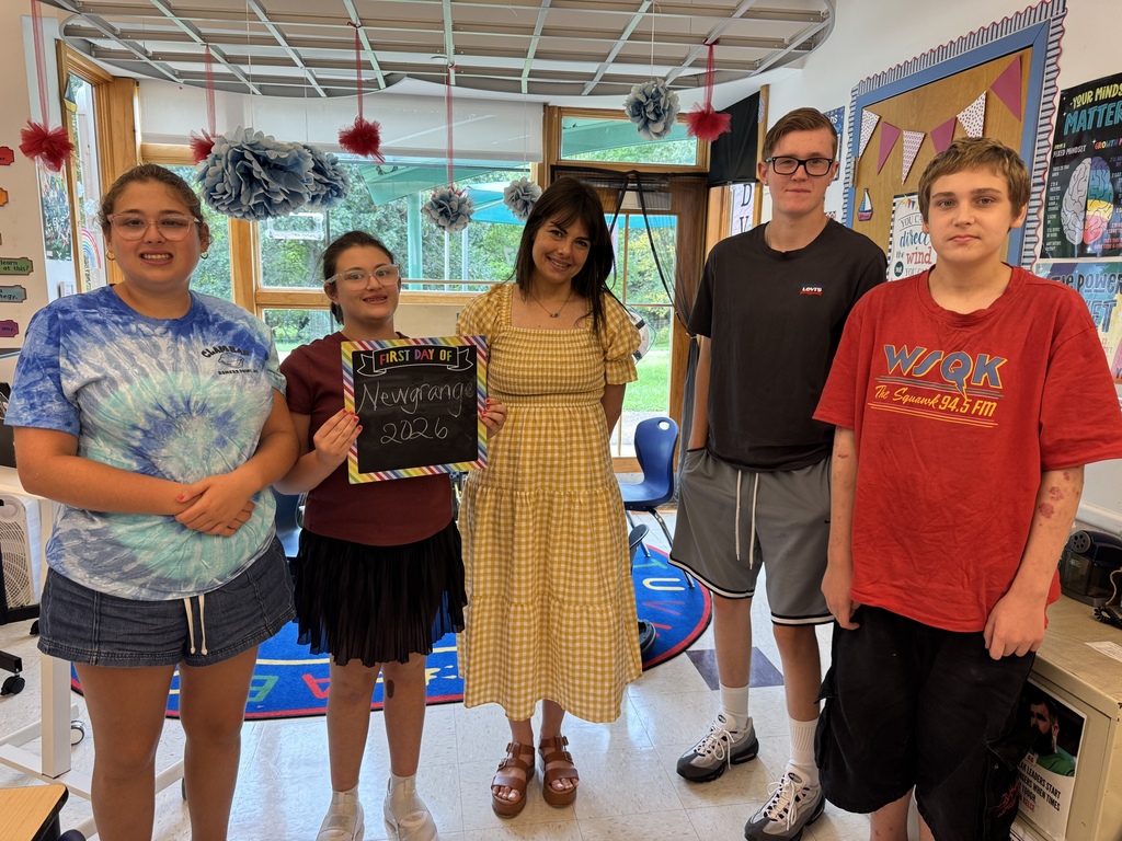 Five students stand together in a classroom with various decorations. A girl in a yellow dress holds a chalkboard.