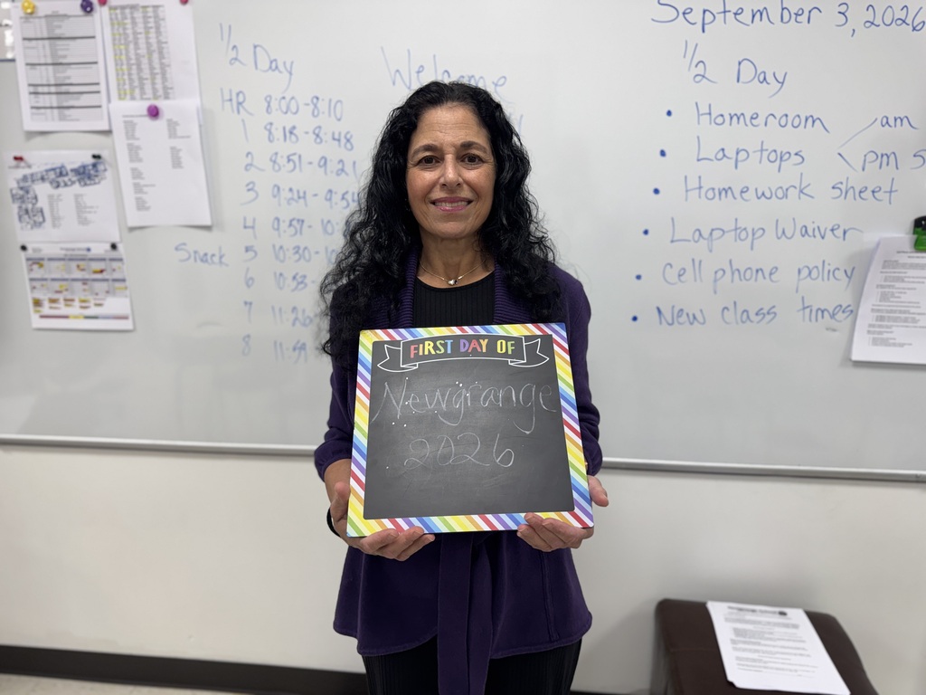 A woman in a classroom, holding a chalkboard reading "Ms. Navarro" and "2020-21." Behind her, a whiteboard lists classroom rules and policies.