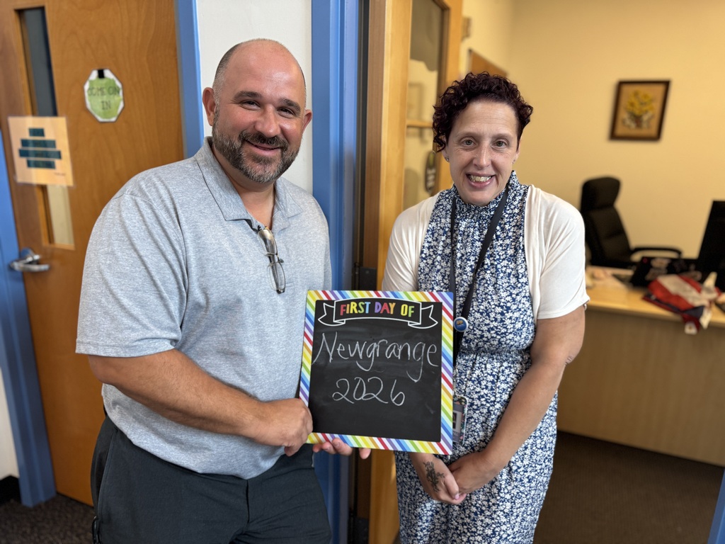 A man and woman stand in an office. He holds a chalkboard with colorful writing. They both smile.