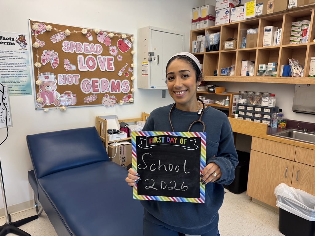 Person in a blue coat holding a chalkboard that says "First Day of School 2026". Behind them is a medical bed and cabinets.