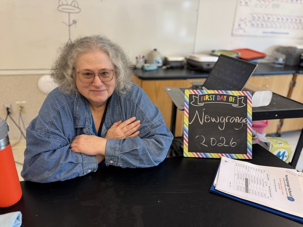 An older woman wearing glasses sits at a desk. Behind her is a counter with items. A blackboard reads "Newyeargang 2026".