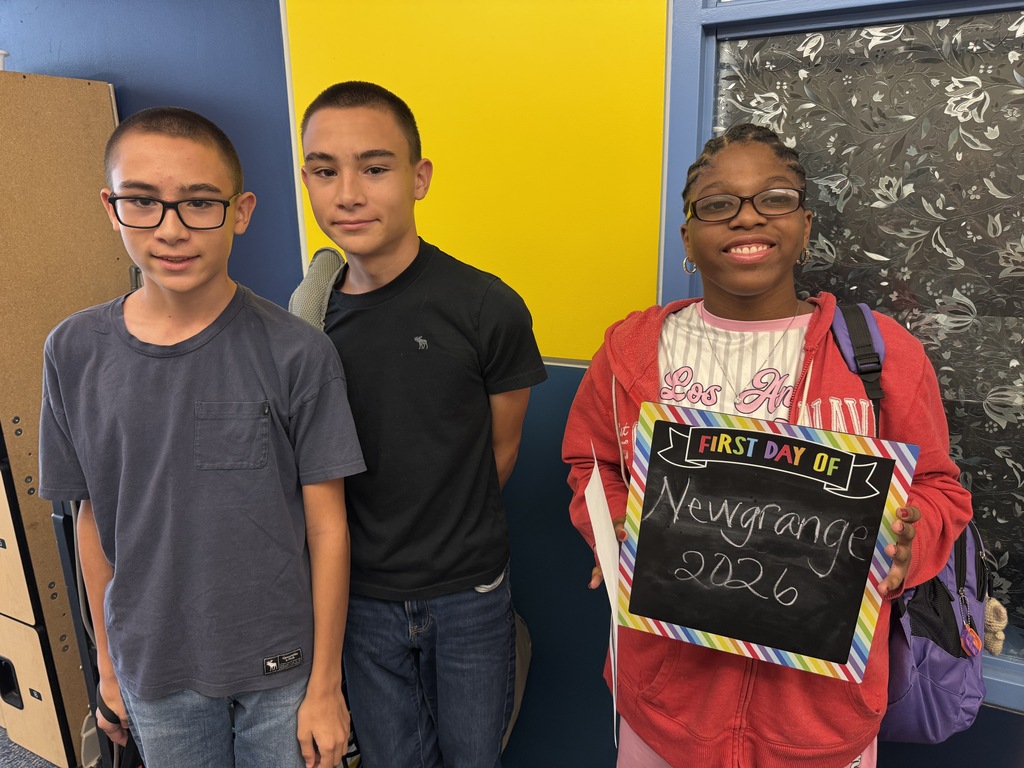 Three students standing next to each other; one holds a sign reading "New Grange 2026". Behind them is a yellow wall with a chalkboard.