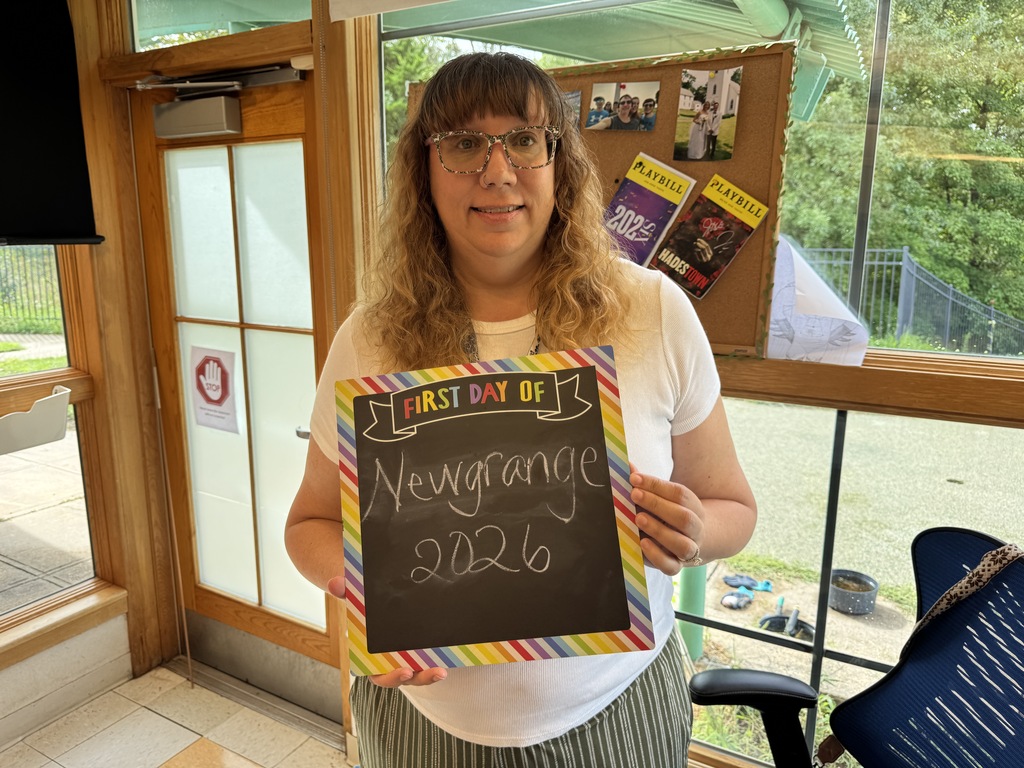 Woman wearing glasses and a white shirt holds up a sign that reads "First Day of New Grange 2026."