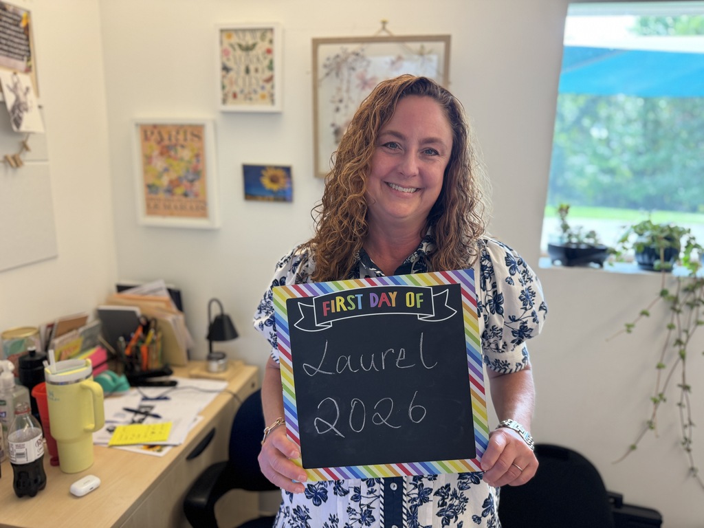 Woman holding a sign that says "First Day of Laurel 2026" standing in a room with white walls and framed art.