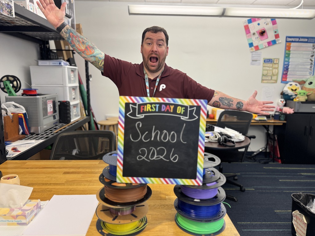 Person in a room with a sign reading "First Day of School 2026" over a stack of spools.