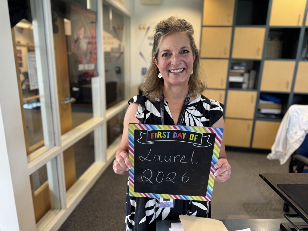 A woman smiles in an office. She holds a chalkboard that reads, "First Day of Laurel 2026."