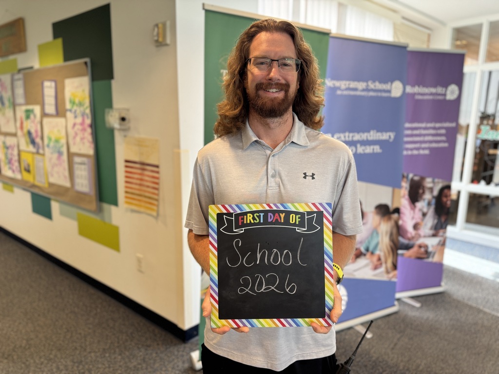 A man in a gray shirt holds a chalkboard with "School 2026" written on it. He is standing in a hallway with colorful posters and banners.