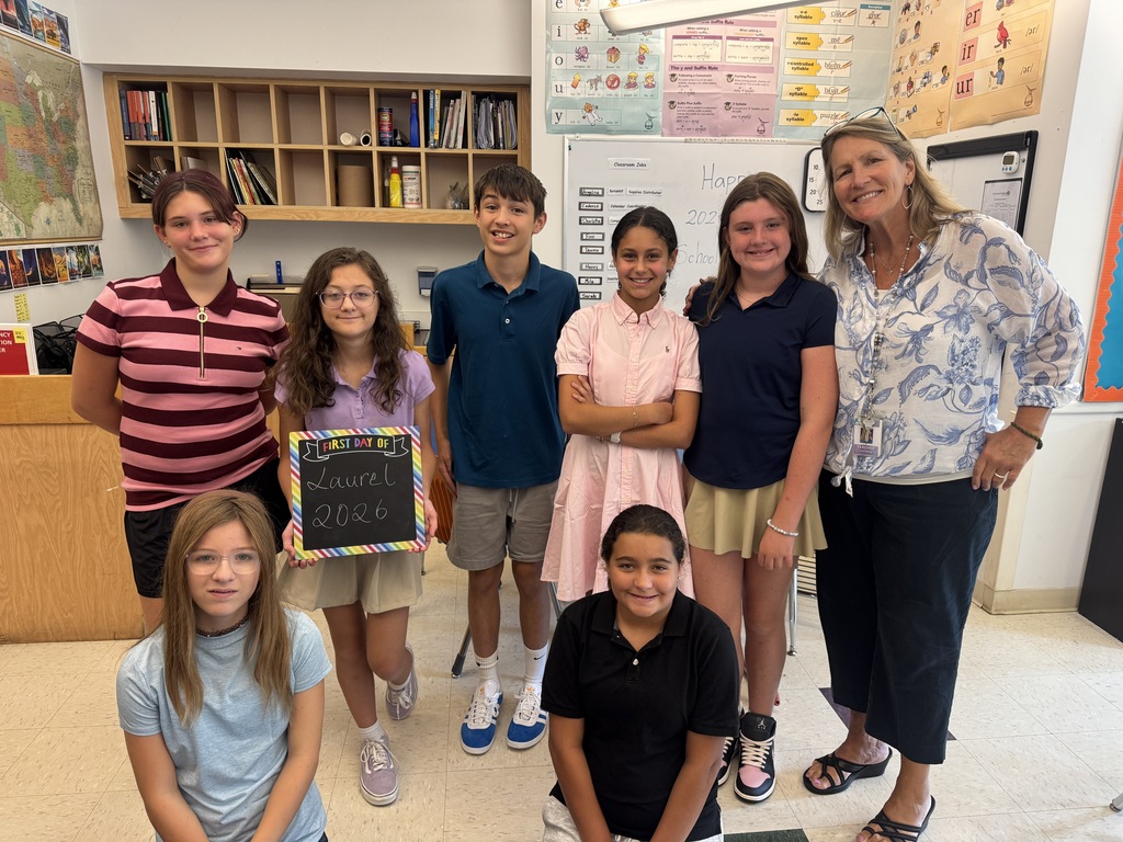 A group of students in a classroom, posing for a photo with a teacher holding a sign.