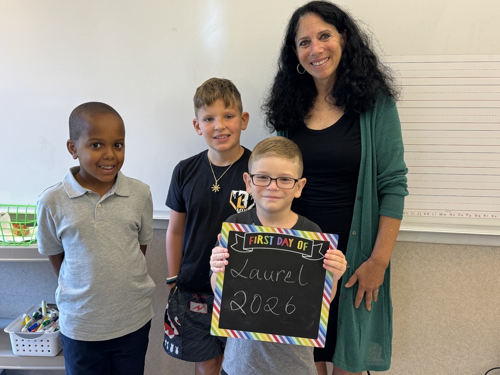 Three children and an adult woman stand in a classroom. One child holds a sign reading "First Day of Laurel 2026".