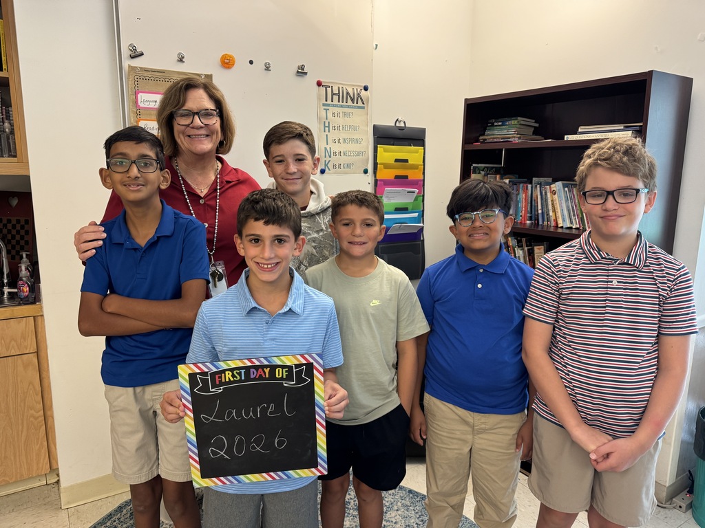 Six boys and a woman stand together, smiling. One boy holds a colorful sign. Behind them, a bookshelf and counter are visible.
