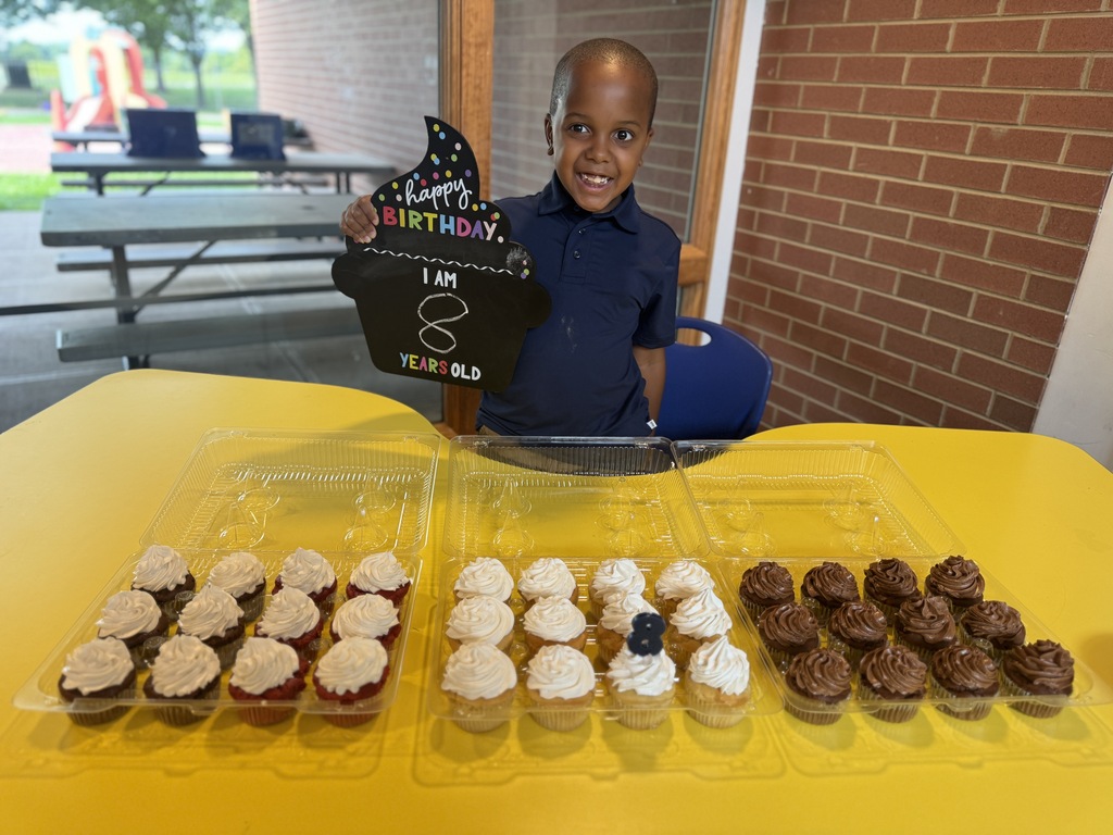 A young boy stands at a table with a "Happy Birthday" sign, displaying multiple rows of cupcakes.