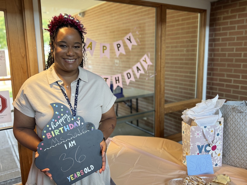 A woman holds a chalkboard that reads "Happy Birthday, I am 36 years old". Behind her is a brick wall, a banner, and a table with gifts.