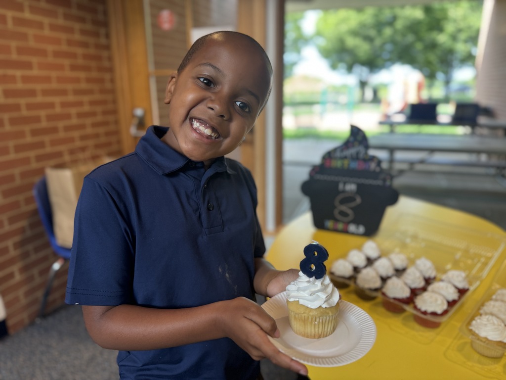 Boy holds a cupcake with "8" written on it in frosting. He is smiling. Cupcake table behind him.
