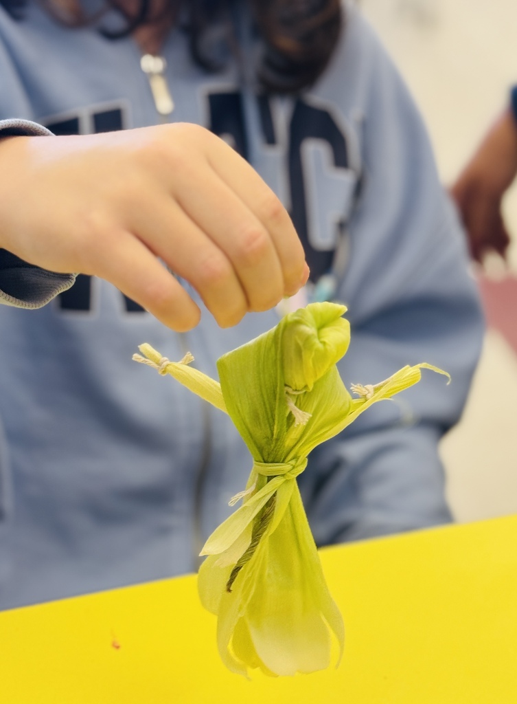 A hand holds a wrapped corn cob on a yellow table. A person in a light blue sweatshirt is in the background.