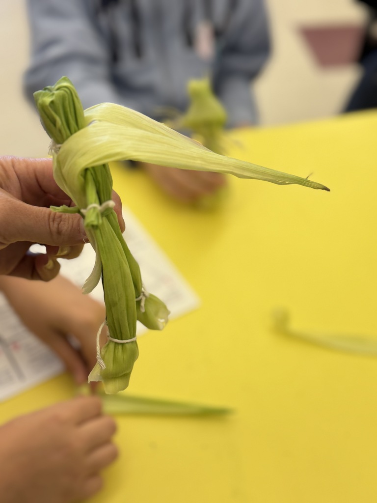 Hands are seen tying corn husks. A yellow table is visible in the foreground.