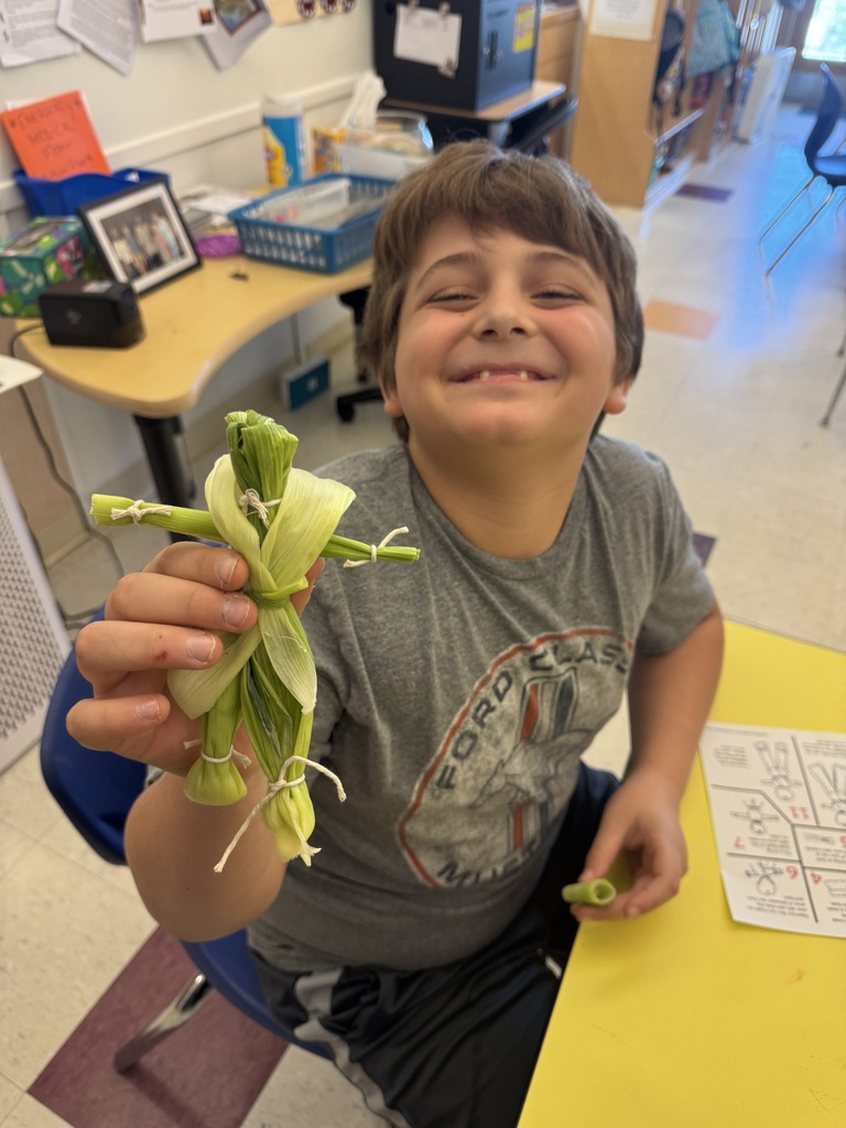 A young boy in a gray t-shirt holds an object made of green beans while smiling. Behind him, a yellow table has papers and a blue chair.