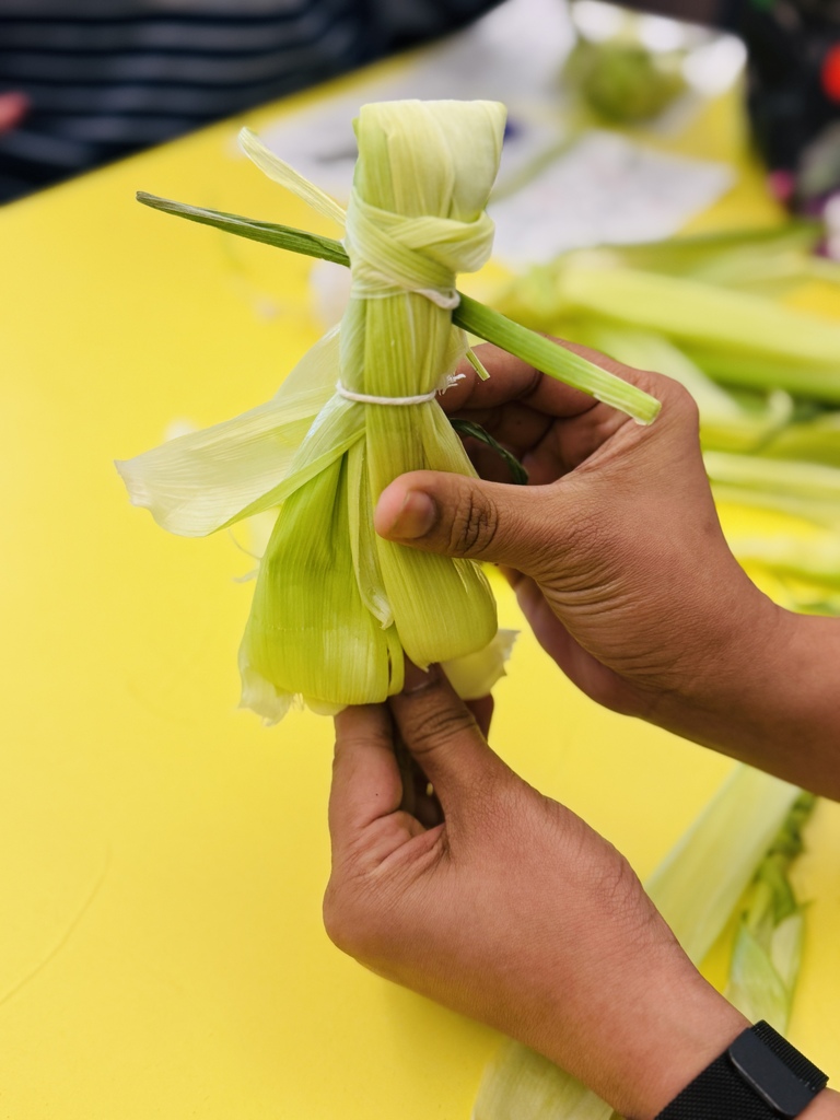 Hands holding a bundle of corn wrapped in husks, set on a yellow table.