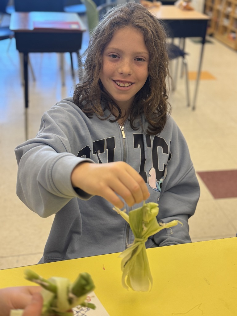 A girl sits at a table with a yellow surface, holding a wrapped plant and smiling.