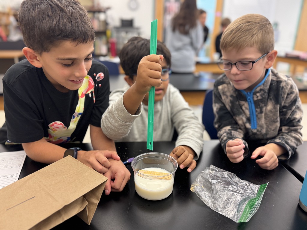 Three boys at a table, one mixing a substance in a cup, another holding a stick, and the last observing.
