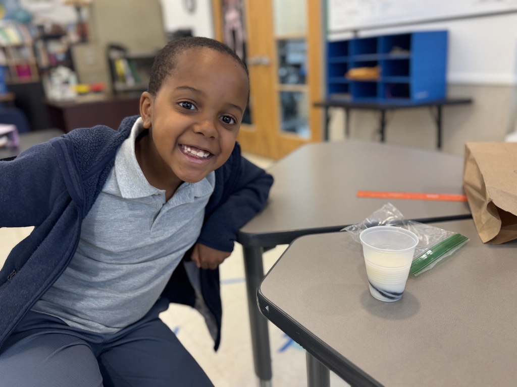 A child smiles at the camera. They sit on the floor in front of a desk. A cup of milk is on the desk.