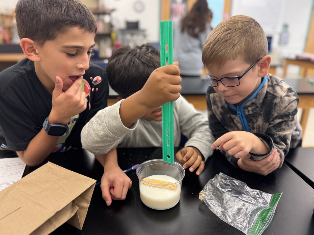 Three kids conduct a science experiment at a lab table, one holding a tube, another tasting milk, and a third observing.