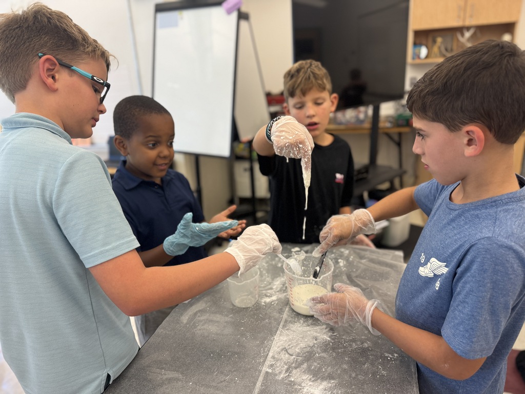 Four children, each wearing gloves, are gathered around a table with science materials in a classroom.