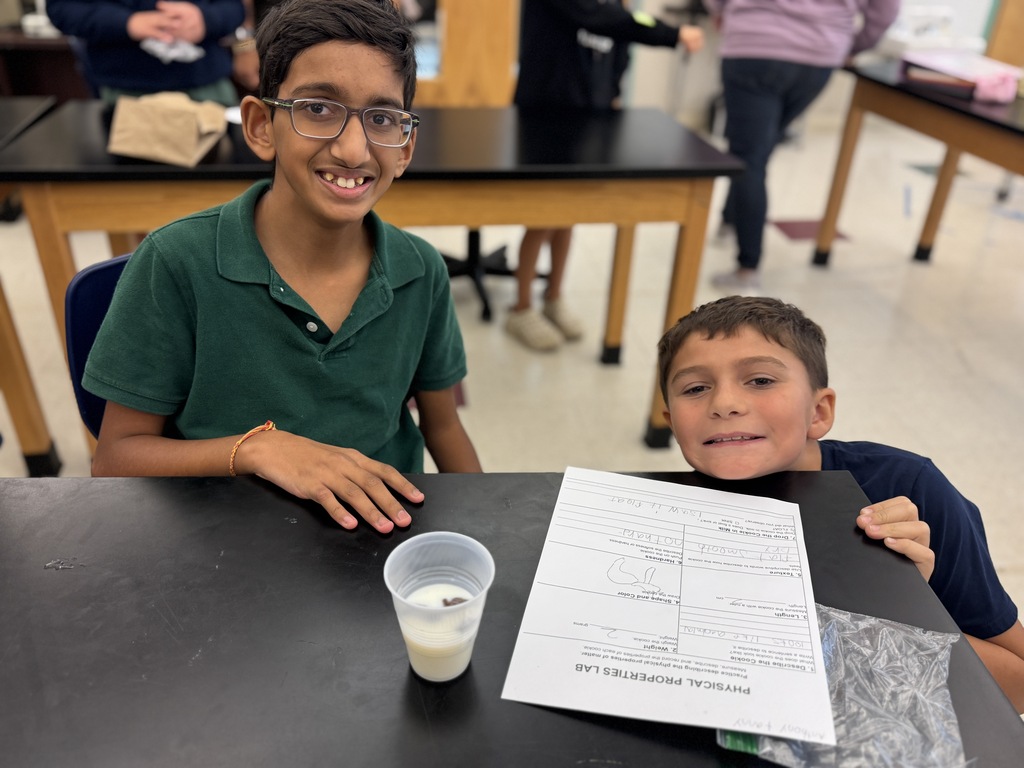 Two boys sit at a table with a cup of milk and a paper in front of them. They sit near tables in a classroom.