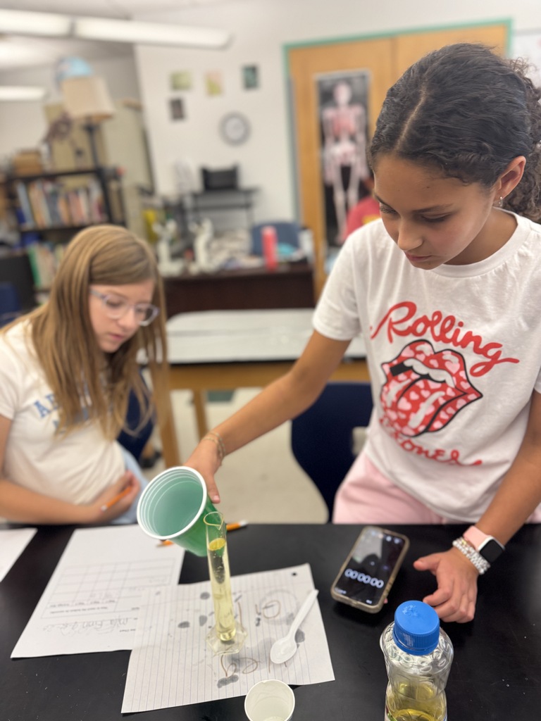 Two girls are seated at a desk, one holding a green cup with a yellow liquid.