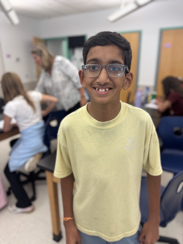 A young boy with glasses and a yellow shirt stands in a classroom, smiling at the camera.