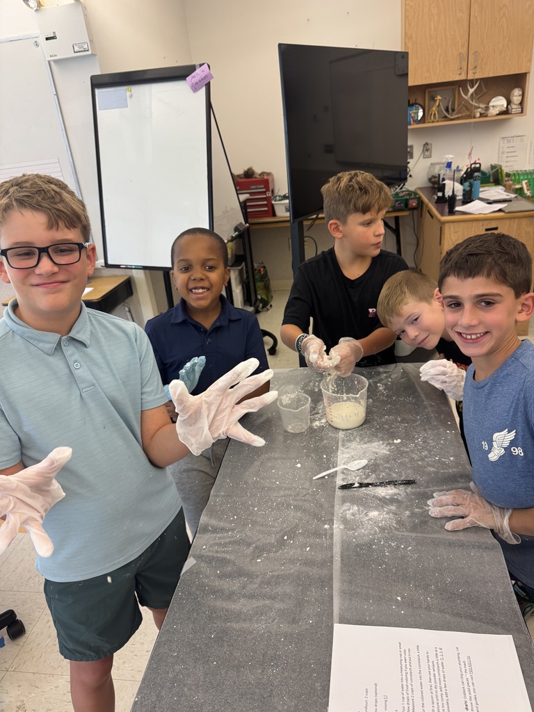 Five children in a classroom, wearing gloves and mixing a substance in a cup. One holds a paper.