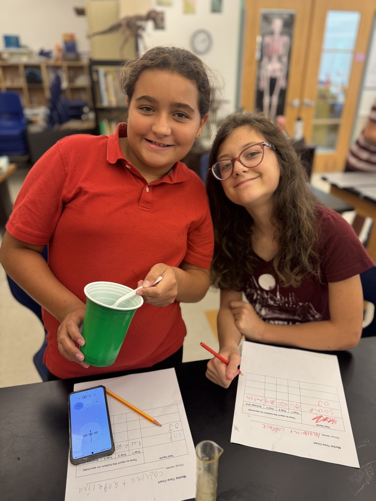 Two young girls in a classroom, one holding a green cup and pen, the other holding a pen and paper.