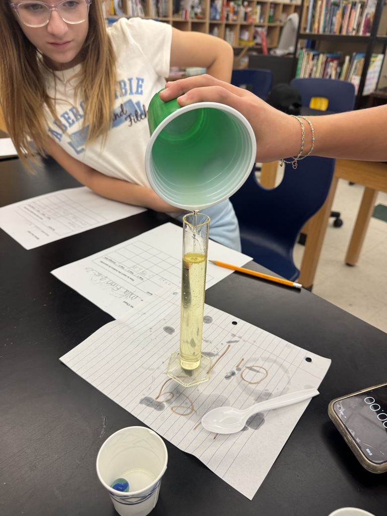 A student pours a yellow liquid from a green cup into a clear test tube.