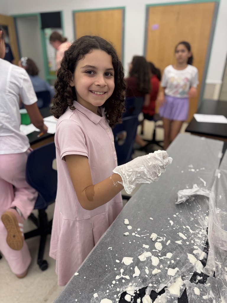 A girl in a pink dress stands at a table, wearing gloves. She has a smile and is surrounded by others in a classroom.
