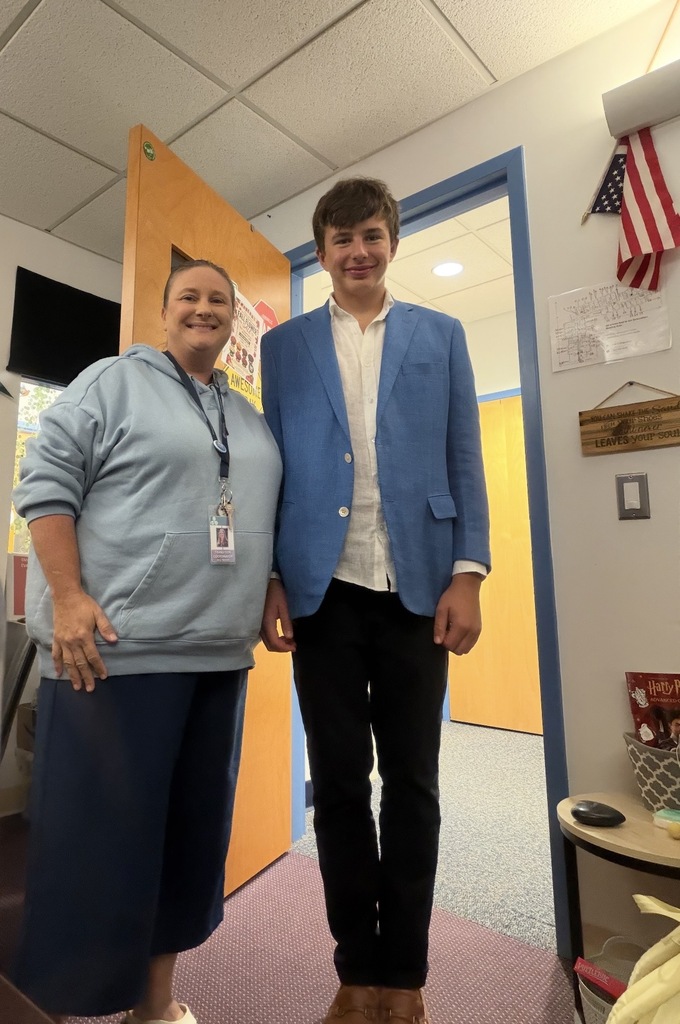 A woman and a man stand in a room with a door. The man wears a blue blazer and black pants.
