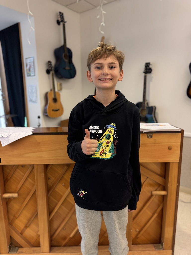 A smiling young boy, wearing a black hoodie, gives a thumbs up in a room with guitars and a wooden counter.