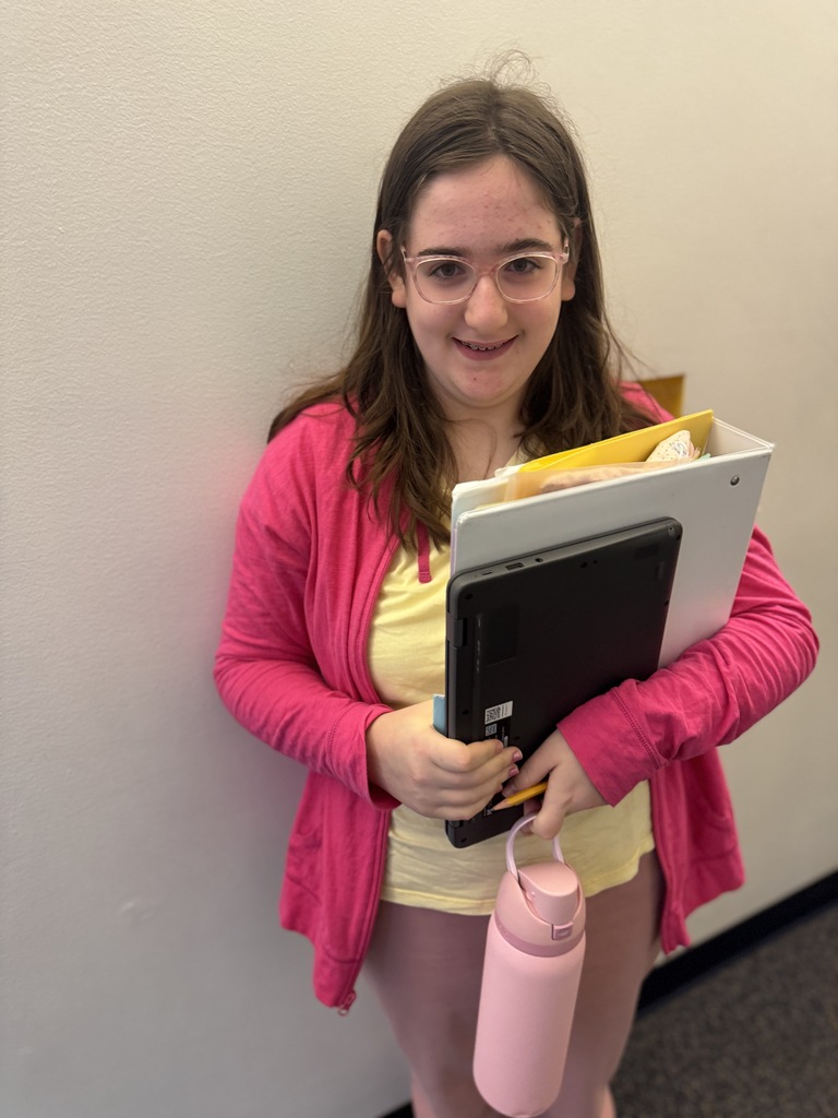 Person in yellow shirt and pink cardigan holding a folder and tablet. Pink water bottle in left hand.