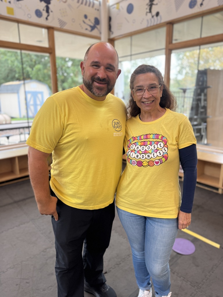 Two individuals, a man and a woman, wearing yellow t-shirts, stand together indoors with windows in the background.